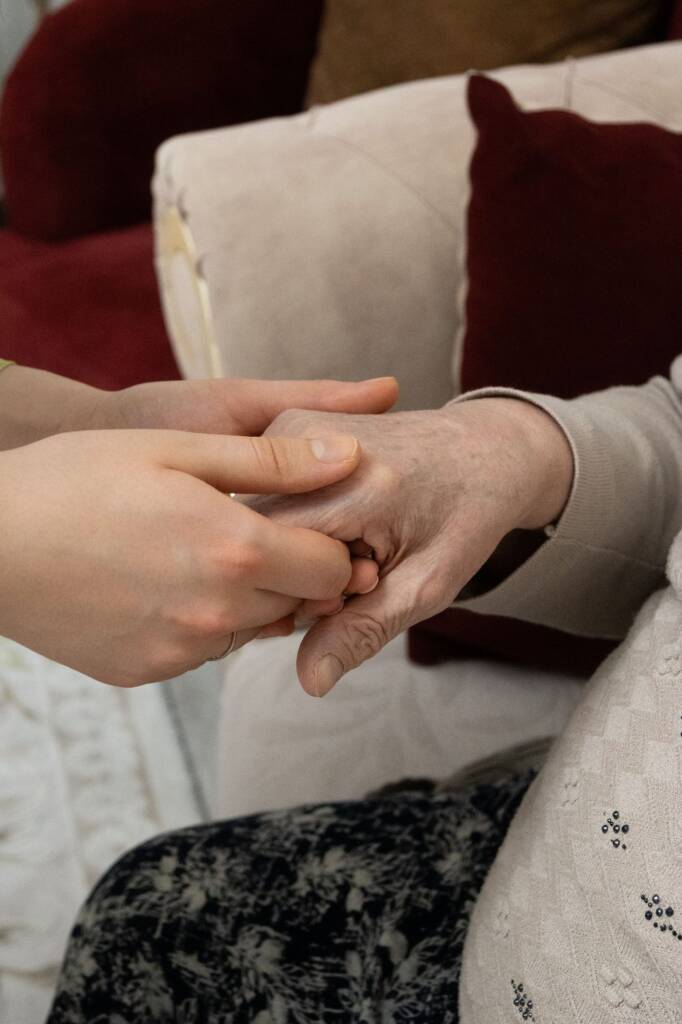 A younger person's hands holding an older person's hand beside an armchair