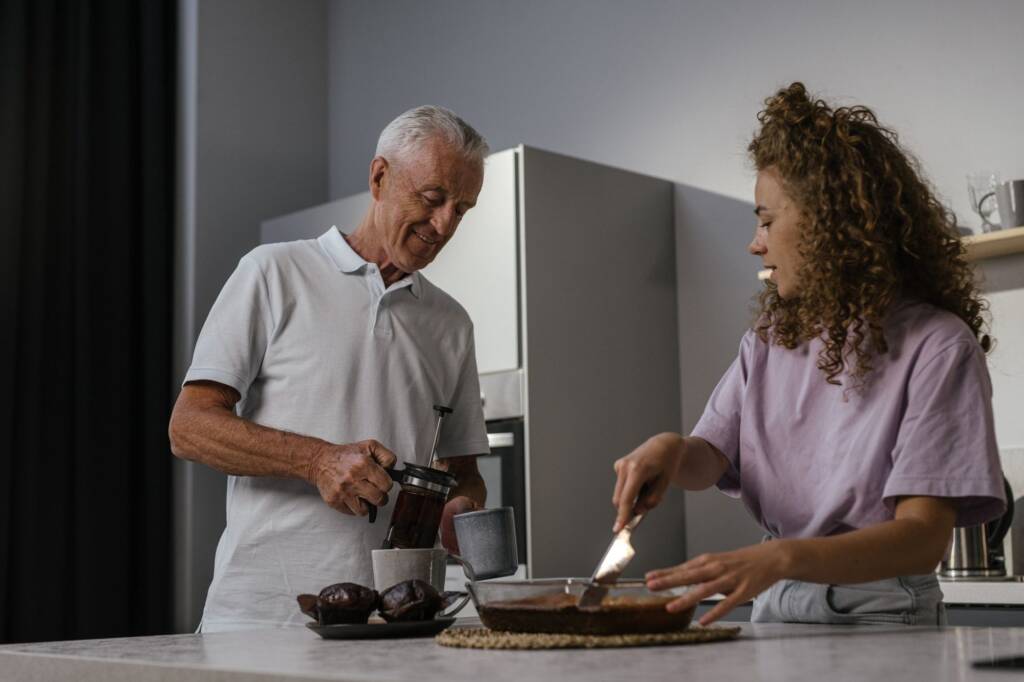 An older man and a younger woman making coffee and cake together in a kitchen
