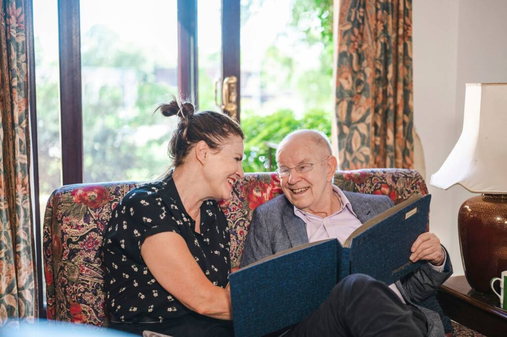 A woman and an older man laughing over a photo album on a sofa