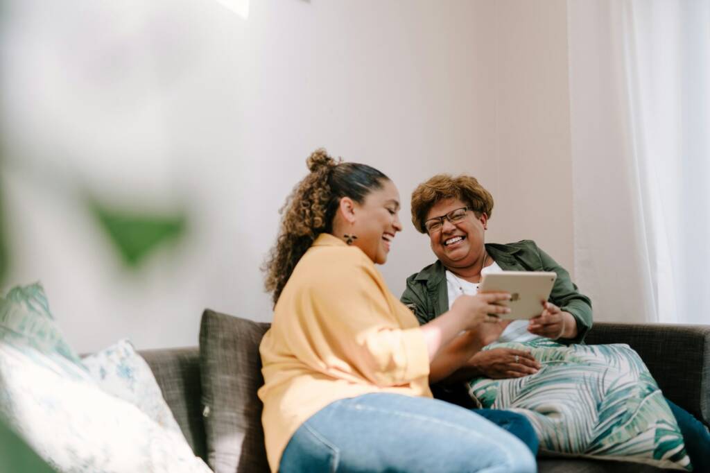 Two women laughing together over a tablet on a sofa at home