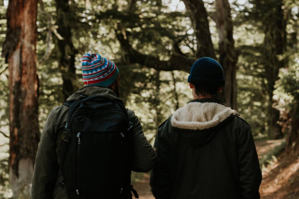 Two young people in woolly hats walking and talking on a woodland path, seen from behind
