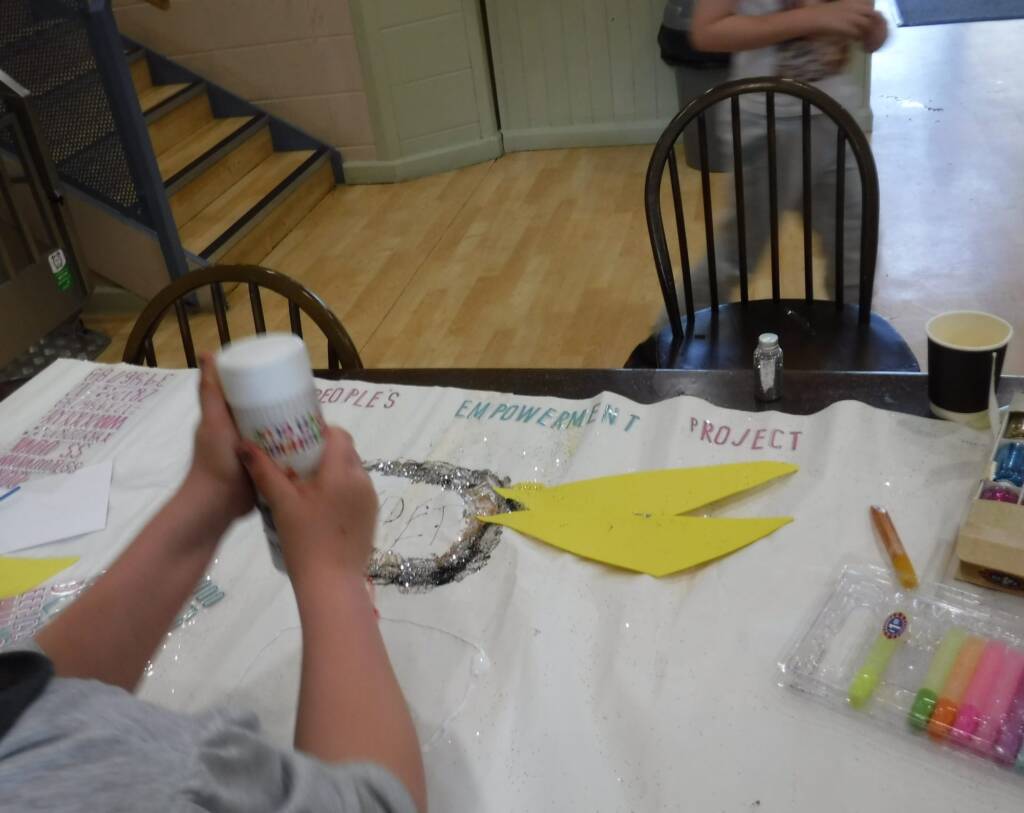 Hands decorating a handmade Young People’s Empowerment Project banner with glitter and card