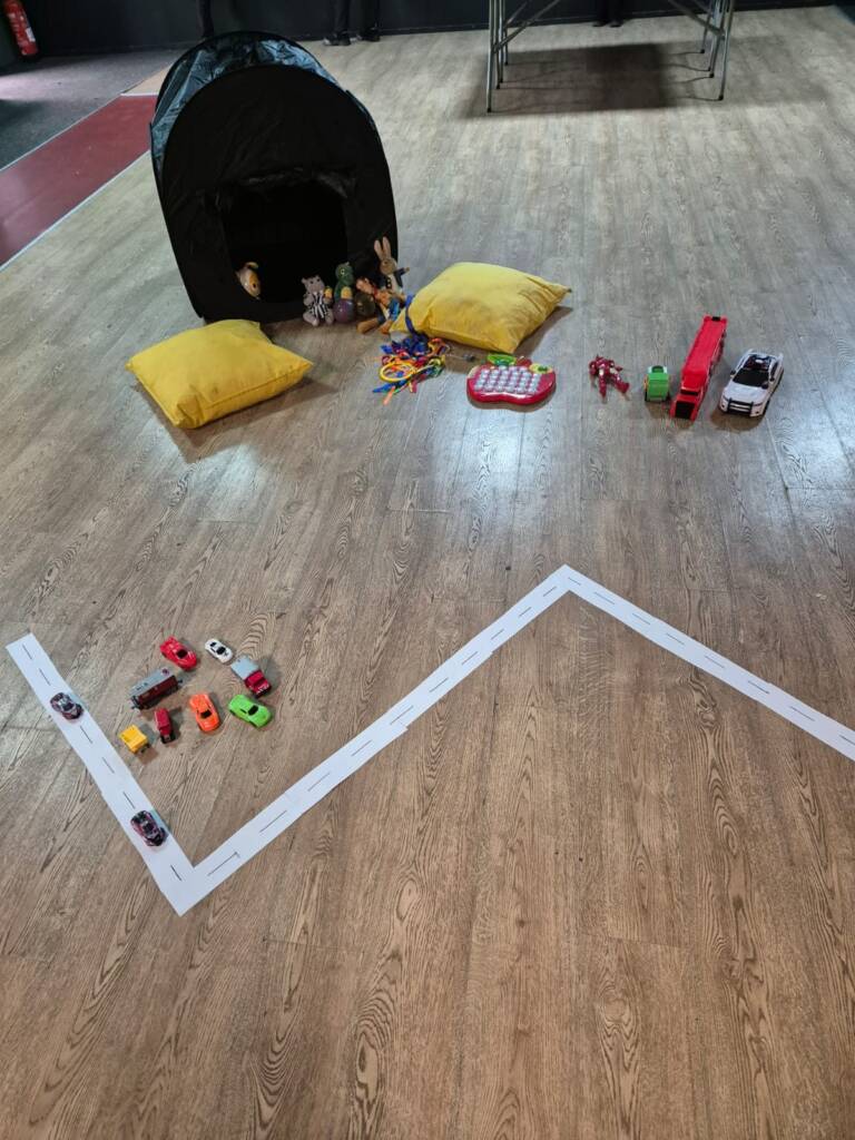 A toy car road marked out in tape on a hall floor, with toy cars, cushions and a play tent